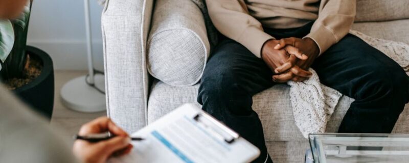 The hands of a Black man are visible, folded as he sits in a therapist’s chair, with the therapist softly out of focus in the background.