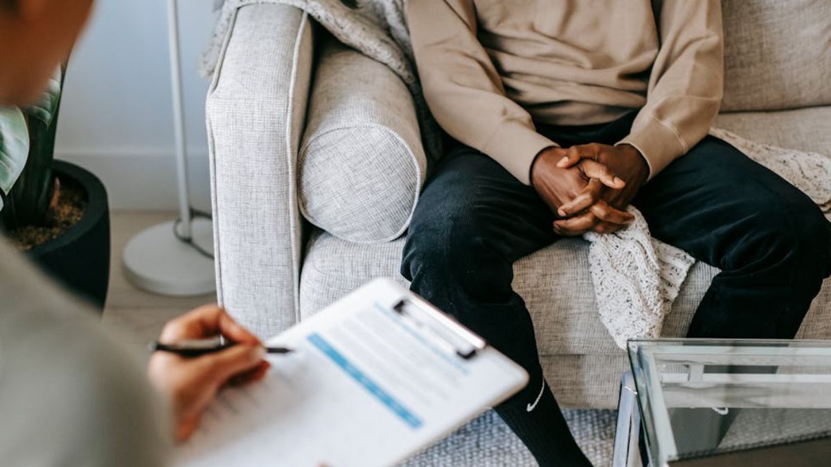 The hands of a Black man are visible, folded as he sits in a therapist’s chair, with the therapist softly out of focus in the background.