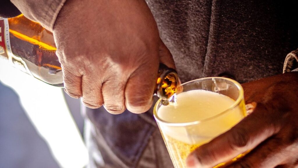 A man’s hand, dressed in a plaid suit sleeve, pours hard liquor into a glass jigger.