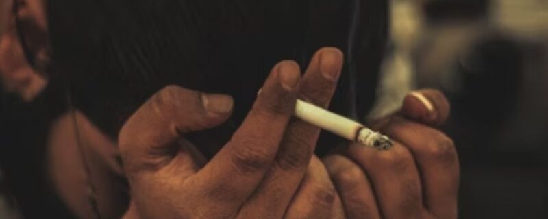 A man holds a cigarette with his face buried in his hands, appearing tired, disturbed, and trapped.