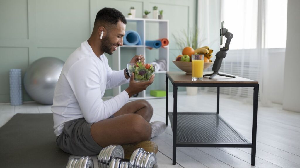A man sits and rests after a workout, with a healthy selection of fruits and vegetables on the table in front of him.