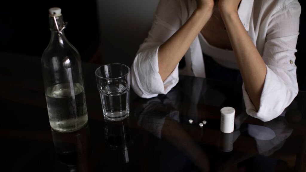 Denial About Substance Use Disorder: A man rests his elbow on a table scattered with bottles of hard liquor and pills, only his torso visible.