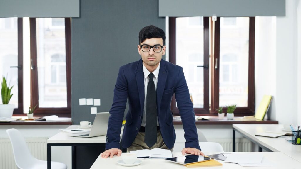 A confident male professional stands at an office table, resting his hands on the surface and looking directly at the camera.