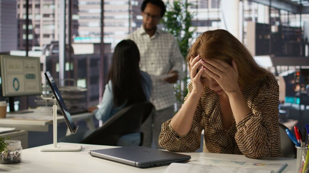 A frustrated female professional rests her elbows on a desk, covering the sides of her forehead with her hands, while coworkers chat in the blurred background.